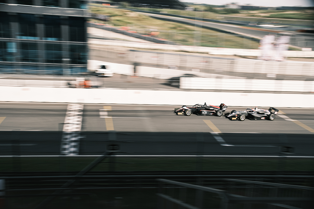 A blurred photo of 2 black single seater race cars approaching the start finish line of a race track