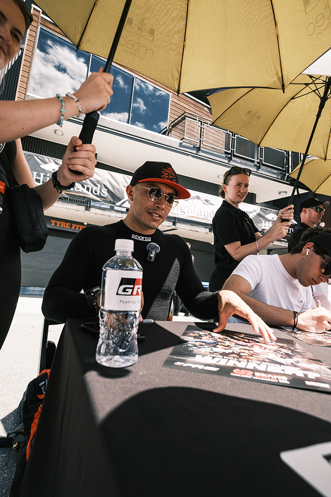 Shawn Rashid sat at a table wearing a red and black cap while an umbrella is held over his head, with a GR branded water bottle in the foreground.